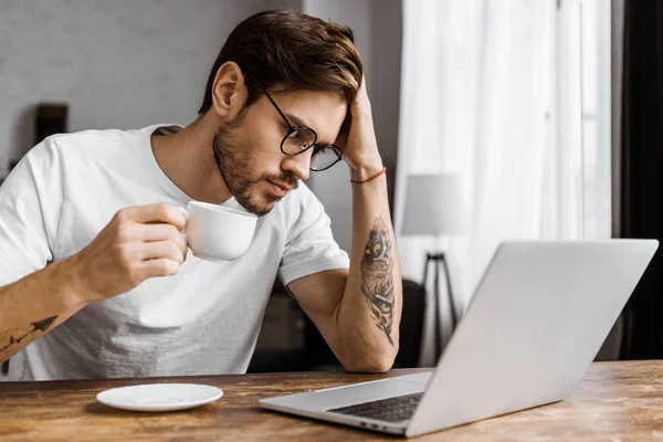 Overworked young freelancer with coffee looking at laptop screen at home — Stock Photo