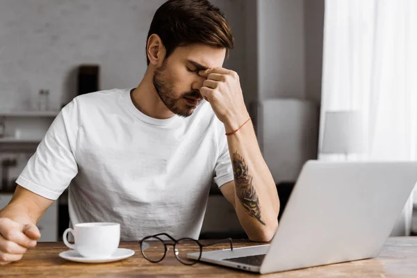 Cansado joven freelancer con café trabajando con el ordenador portátil en casa - foto de stock