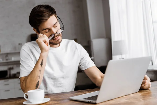 Somnolent jeune pigiste avec café travaillant avec ordinateur portable à la maison — Photo de stock