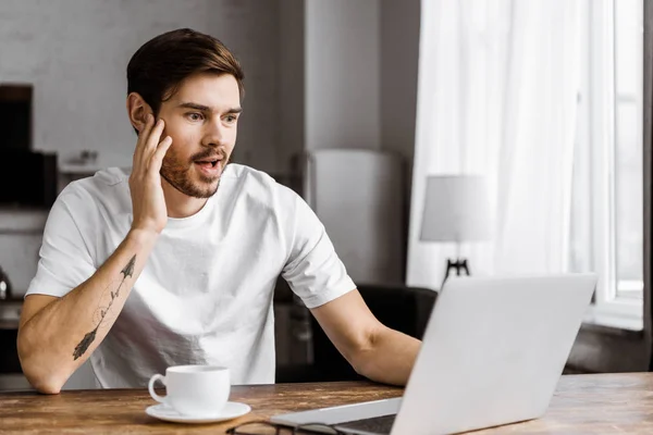 Surprised young man with coffee using laptop at home — Stock Photo