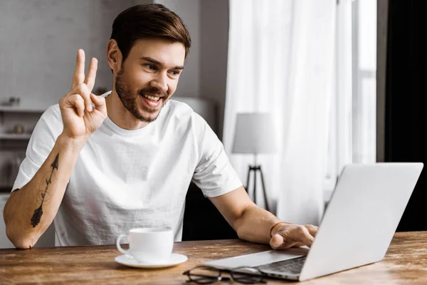 Handsome young man with coffee making video call with laptop and making peace gesture at home — Stock Photo