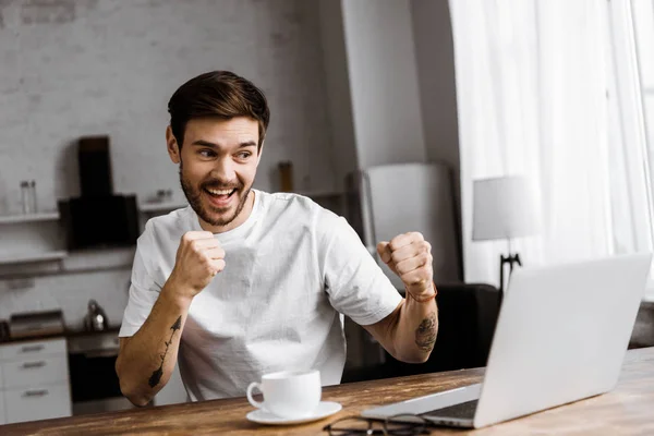 Beau jeune homme avec café faire appel vidéo avec ordinateur portable et montrant les poings à la maison — Photo de stock