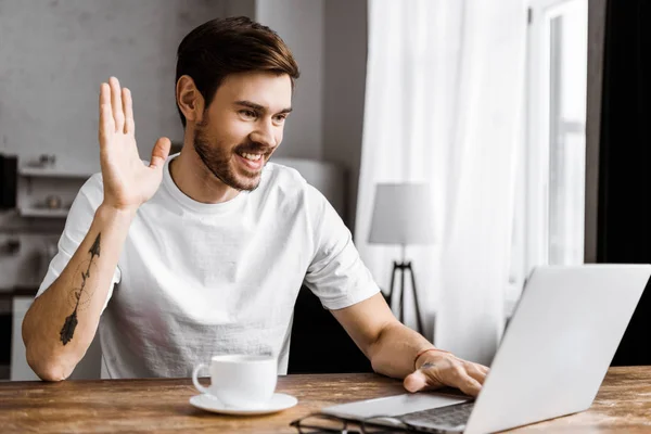 Handsome young man with coffee making video call with laptop and waving at home — Stock Photo