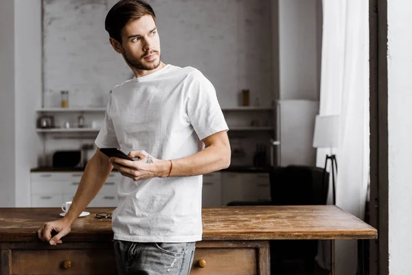 Bello giovane utilizzando smartphone e guardando lontano in cucina a casa — Foto stock