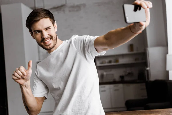 Bonito jovem tomando selfie e fazendo polegar até gesto em casa — Fotografia de Stock