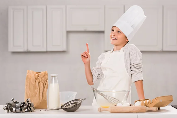 Веселый мальчик в шляпе шеф-повара и фартуке делает жест идеи во время приготовления пищи за столом на кухне — стоковое фото