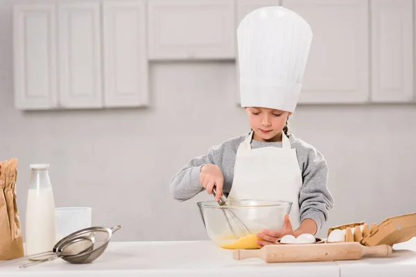 Niño concentrado en el sombrero del chef batiendo huevos en un tazón en la mesa en la cocina - foto de stock