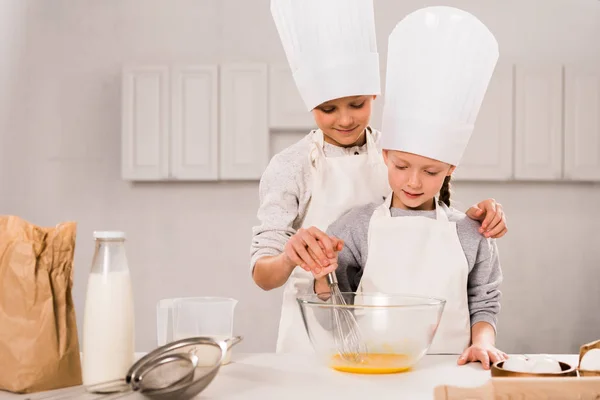 Enfants dans des tabliers et des chapeaux de chef fouettant des œufs dans un bol à table dans la cuisine — Photo de stock