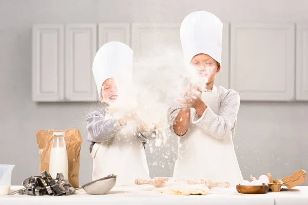 Niños felices en sombreros de chef divirtiéndose con harina en la cocina - foto de stock