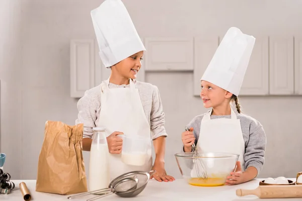 Niño pequeño con leche y hermana mirándose cerca de la mesa en la cocina - foto de stock