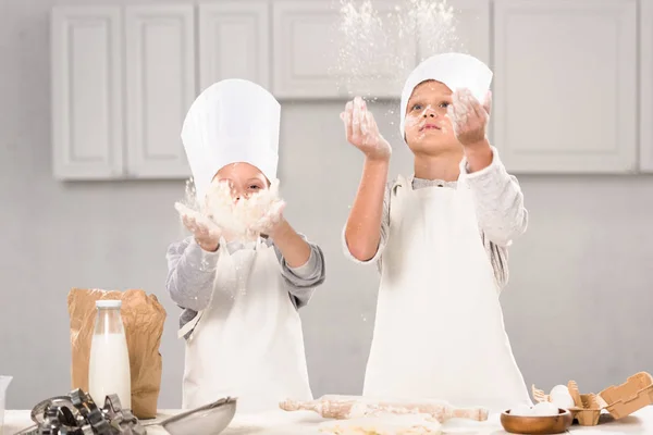 Сестра и брат в шляпах шеф-повара веселятся с мукой на кухне — стоковое фото