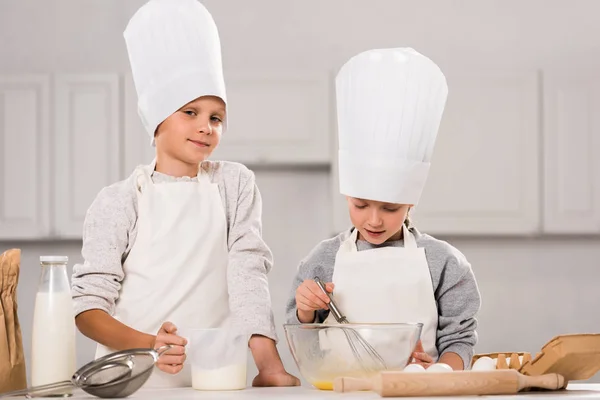 Kleiner Junge blickt in die Kamera und seine Schwester verquirlt Eier in Schüssel am Tisch in der Küche — Stockfoto