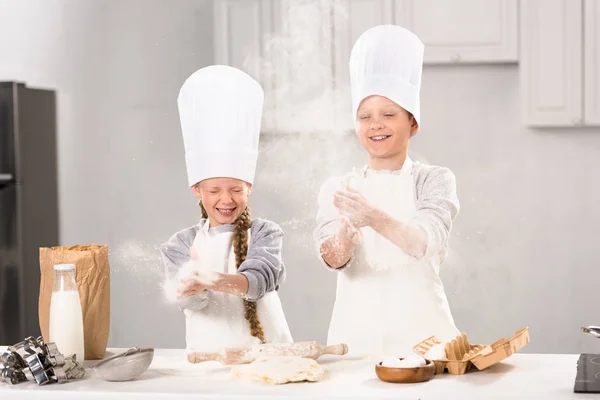 Селективное внимание счастливых детей в шляпах шеф-повара, веселящихся с мукой на кухне — стоковое фото