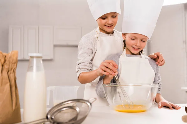 Boy with sister in chef hats whisking eggs in bowl at table in kitchen — Stock Photo