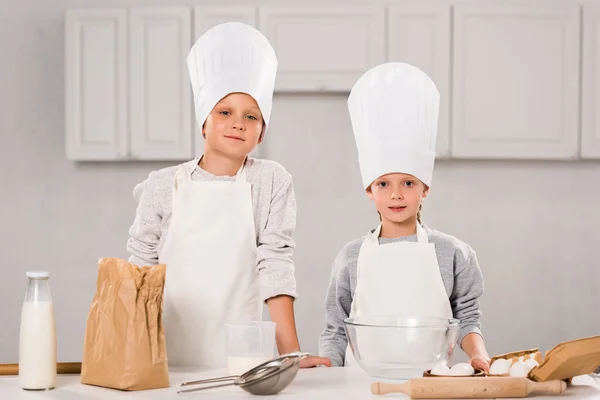 Hermano y hermana en delantales y sombreros de chef mirando a la cámara en la cocina - foto de stock