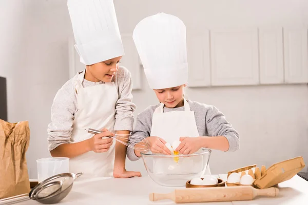 Enfoque selectivo de en sombreros de chef y delantales batiendo huevos en tazón en la mesa en la cocina - foto de stock