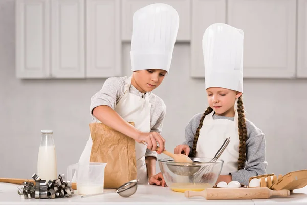 Brother and sister in chef hats and aprons whisking eggs in bowl at table in kitchen — Stock Photo