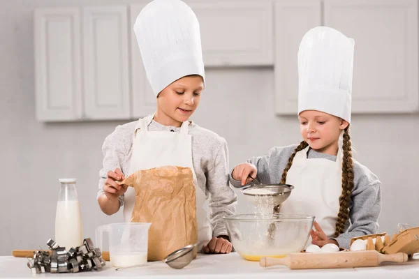 Щасливі діти в фартухах, що просіюють борошно через сито в миску за столом на кухні — стокове фото