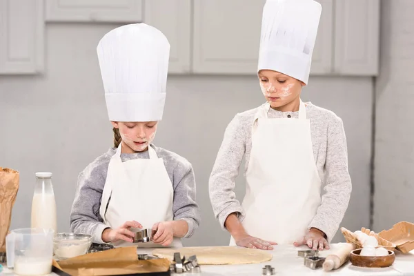 Schwester und Bruder in Kochmützen und Schürzen schneiden Teig für Plätzchen am Tisch in der Küche aus — Stockfoto