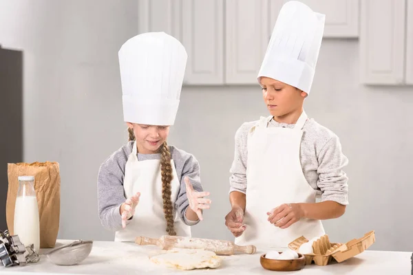 Petits enfants dans des tabliers et des chapeaux de chef faire de la pâte avec rouleau à pâtisserie à table dans la cuisine — Photo de stock