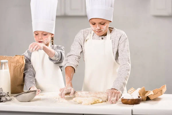 Селективное внимание брата и сестры в фартуках и шляпах шеф-повара, делающих тесто со скалкой за столом на кухне — стоковое фото