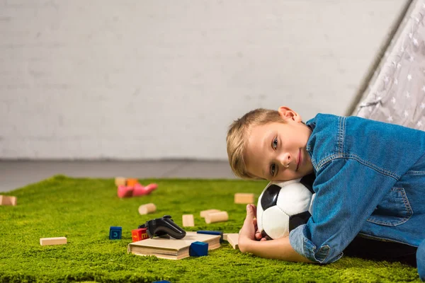 Чарівний хлопчик лежить з футбольним м'ячем на зеленому газоні вдома — стокове фото