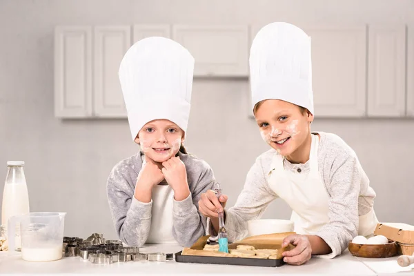 Enfants joyeux dans des tabliers brossant des biscuits sur une plaque de cuisson dans la cuisine — Photo de stock