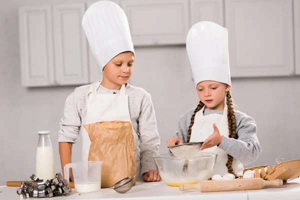 Sorella e fratello in grembiuli setacciando farina attraverso setaccio in ciotola a tavola in cucina — Foto stock