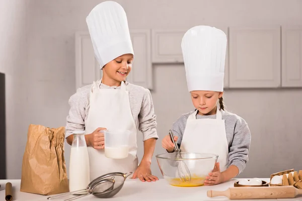 Brother and sister in chef hats and aprons whisking eggs in bowl at table in kitchen — Stock Photo