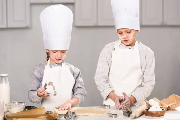 Crianças em chapéus de chef e aventais cortando a massa de farinha de biscoitos à mesa na cozinha — Fotografia de Stock