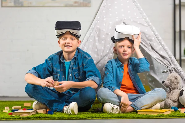 Happy kids with virtual reality headsets looking at camera and sitting near wigwam at home — Stock Photo