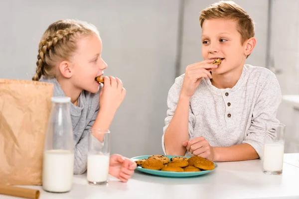 Crianças felizes comendo biscoitos e bebendo leite à mesa na cozinha — Fotografia de Stock