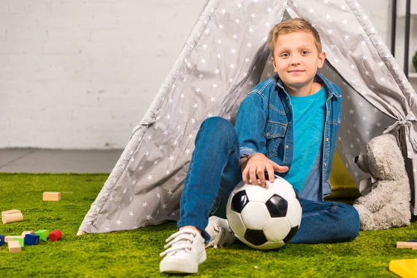 Positiver kleiner Junge sitzt mit Fußballball in der Nähe von Zelt zu Hause — Stockfoto