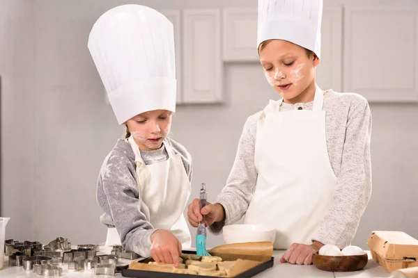 Концентрированные дети в фартуках чистят печенье на подносе для выпечки на кухне — стоковое фото