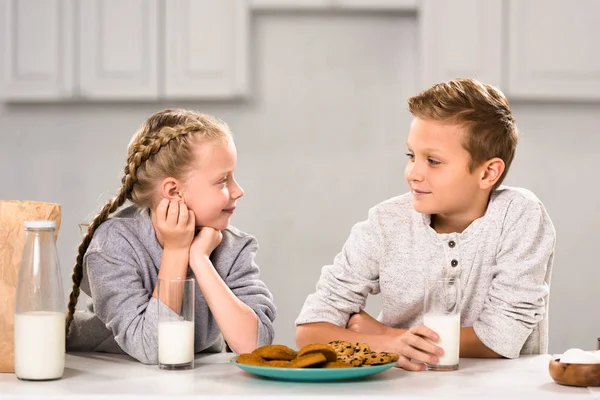 Улыбающиеся дети смотрят друг на друга и сидят за столом с печеньем и молоком на кухне — стоковое фото