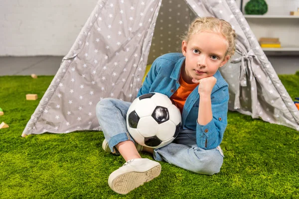 Selective focus of kid with soccer ball looking at camera while sitting on green lawn near tent at home — Stock Photo