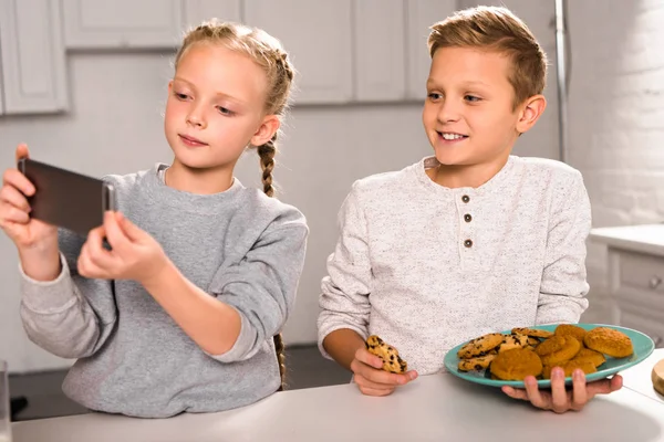 Очаровательный ребенок делает селфи на смартфоне с братом держит тарелку с вкусным печеньем на кухне — стоковое фото