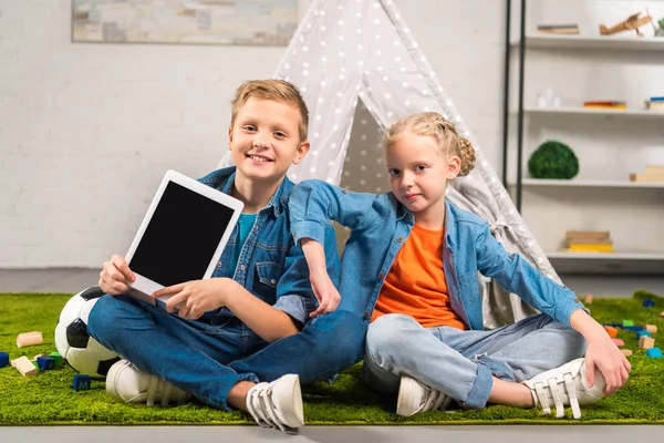 Niños pequeños y felices mostrando tableta digital con pantalla en blanco cerca de la tienda en casa - foto de stock