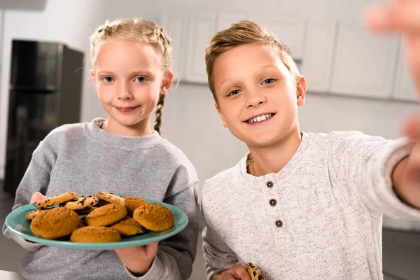 Selektiver Fokus des Jungen, der Selfie mit Schwester macht, während sie Teller mit Keksen hält — Stockfoto