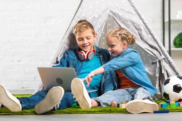 Bambino che punta sullo schermo del computer portatile per sorridere fratello con le cuffie sopra il collo vicino alla tenda a casa — Foto stock