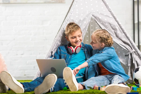 Enfant pointant vers l'écran d'ordinateur portable pour sourire frère avec des écouteurs sur le cou près de la tente à la maison — Photo de stock