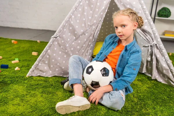 Criança com bola de futebol olhando para a câmera enquanto sentado no gramado verde perto da barraca em casa — Fotografia de Stock
