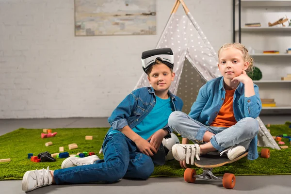 Маленький мальчик с наушниками виртуальной реальности над головой и его сестра сидит на скейтборде рядом с wigwam дома — стоковое фото