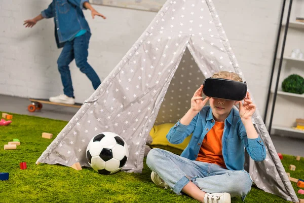 Kid using virtual reality headset near wigwam while her brother riding on skateboard behind at home — Stock Photo