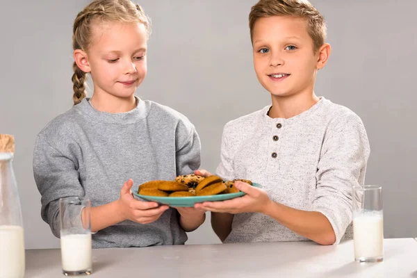 Happy brother and sister holding plate with cookies near table with milk glasses — Stock Photo