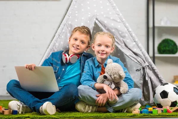 Bambini gioiosi con computer portatile guardando la fotocamera e seduti con tenda a casa — Foto stock