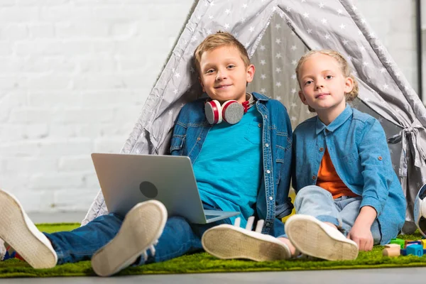 Niños pequeños felices con el ordenador portátil mirando a la cámara y sentado con la tienda de campaña en casa - foto de stock