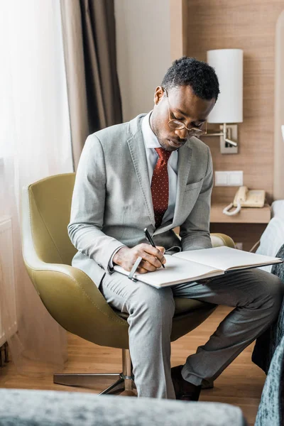 Guapo hombre de negocios afroamericano en traje gris escribiendo en diario en habitación de hotel - foto de stock