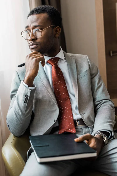 Homme d'affaires afro-américain réfléchi en costume gris assis dans un fauteuil avec journal dans la chambre d'hôtel — Photo de stock