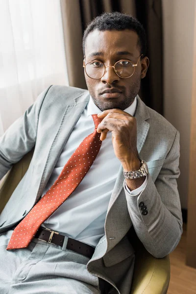 Pensativo hombre de negocios afroamericano en traje gris y corbata roja sentado en sillón - foto de stock